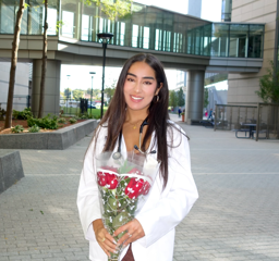  A full-body outdoor photo of Mitra standing in a courtyard. She is smiling at the camera and wearing a white coat over her clothing. She holds a bouquet of red roses in front of her.