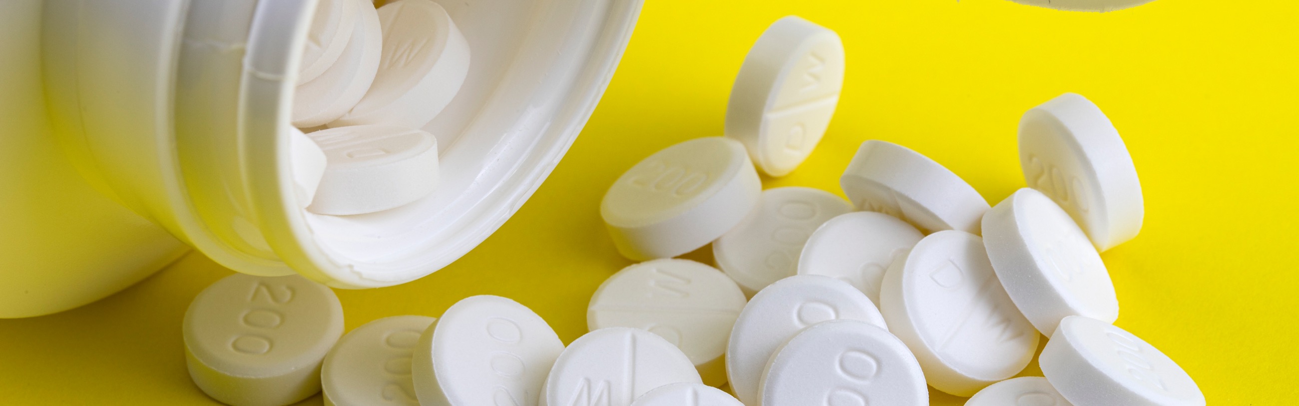 Round white pills spill out of a bottle onto a yellow background